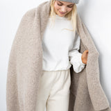 JONES + CO oatmeal alpaca wool blend throw blanket draped over a woman wearing a grey beanie and white sweater.