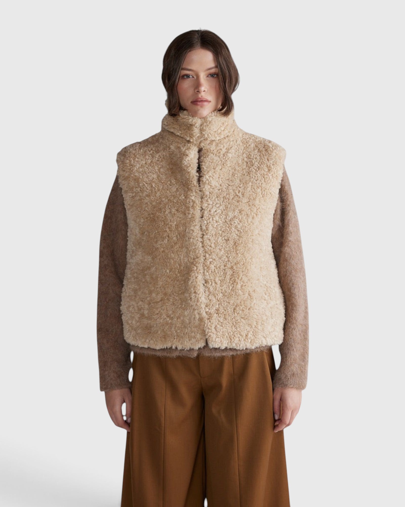 JONES + CO oatmeal high neck reversible faux fur vest with a cropped, boxy silhouette worn over a brown knit top.