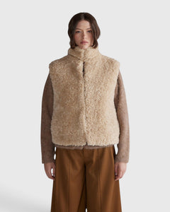 JONES + CO oatmeal high neck reversible faux fur vest with a cropped, boxy silhouette worn over a brown knit top.