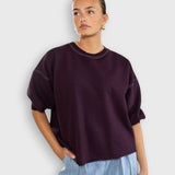 Model wearing a JONES + CO plum crewneck sweatshirt with drop shoulders, cropped sleeves, and raw edge hemline.