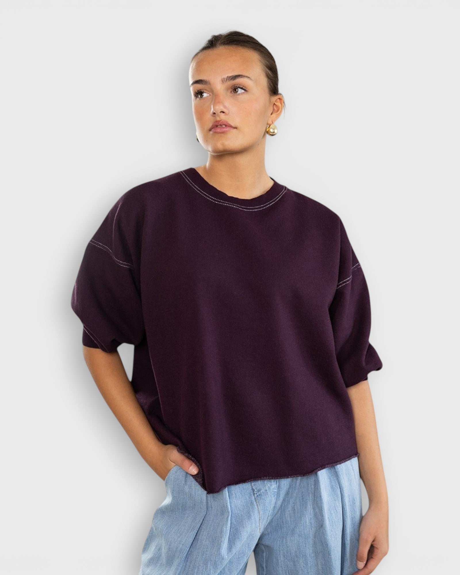 Model wearing a JONES + CO plum crewneck sweatshirt with drop shoulders, cropped sleeves, and raw edge hemline.