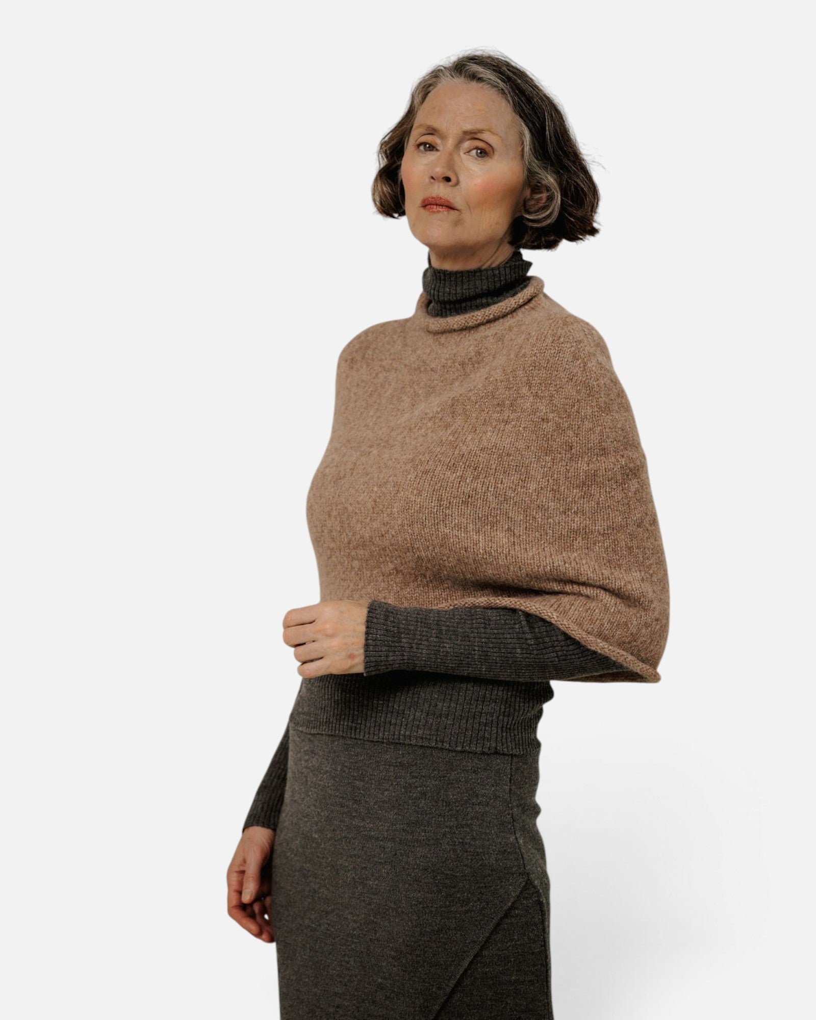 JONES + CO seamless alpaca-blend poncho in dark fawn worn over a charcoal turtleneck sweater by a mature woman.