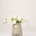 JONES + CO tall stoneware vase with a beige glaze and brown drip detail, holding white ranunculus flowers.