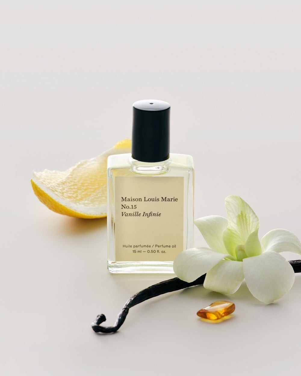 JONES + CO No.15 Vanille Infinie perfume oil in a clear glass bottle with a black cap, next to a lemon slice, vanilla bean, and orchid.
