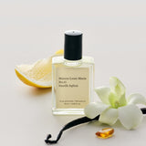 JONES + CO No.15 Vanille Infinie perfume oil in a clear glass bottle with a black cap, next to a lemon slice, vanilla bean, and orchid.