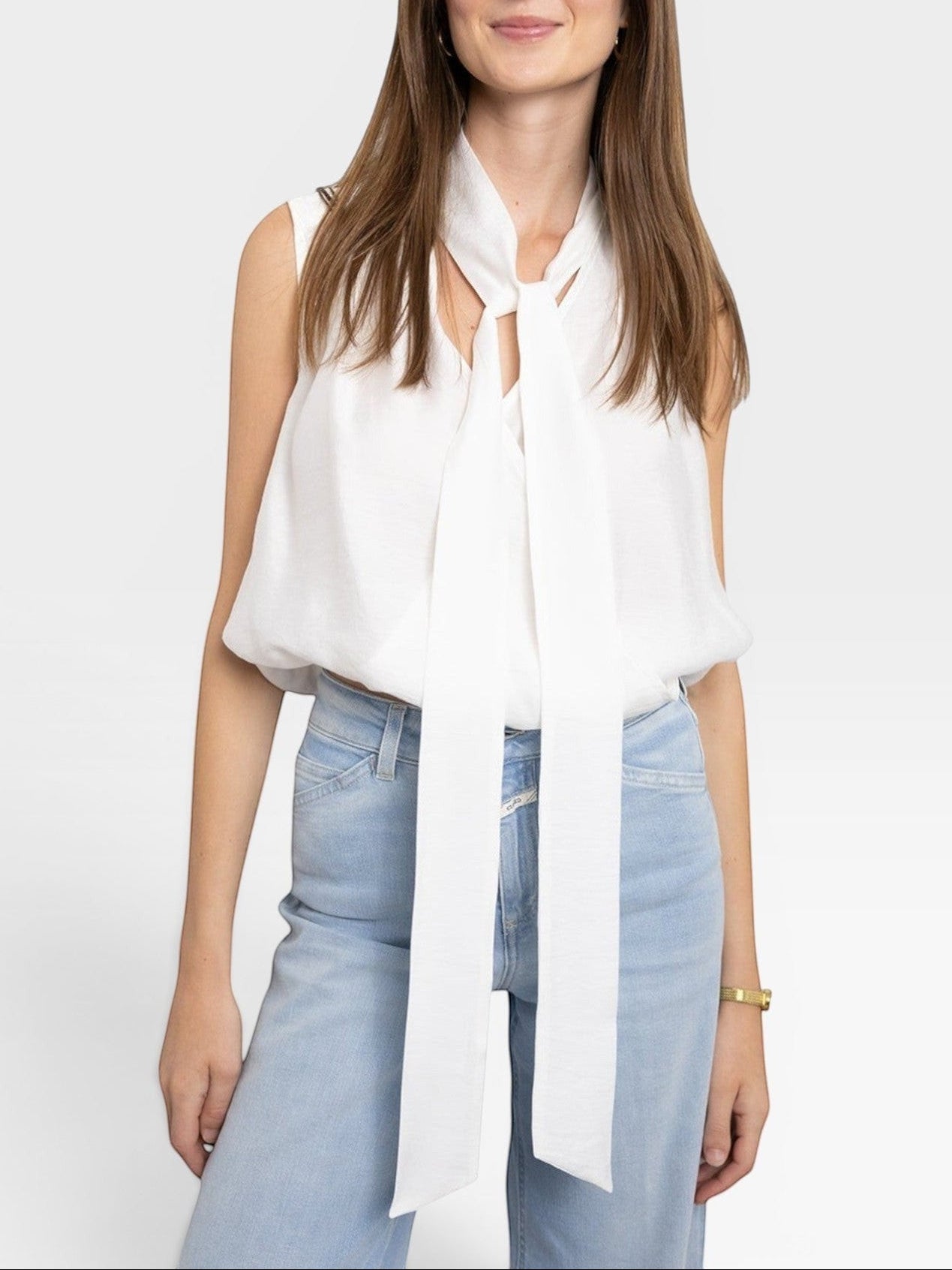 JONES + CO white sleeveless blouse with tie-neck detail, paired with light wash jeans. Model is wearing a gold bracelet.
