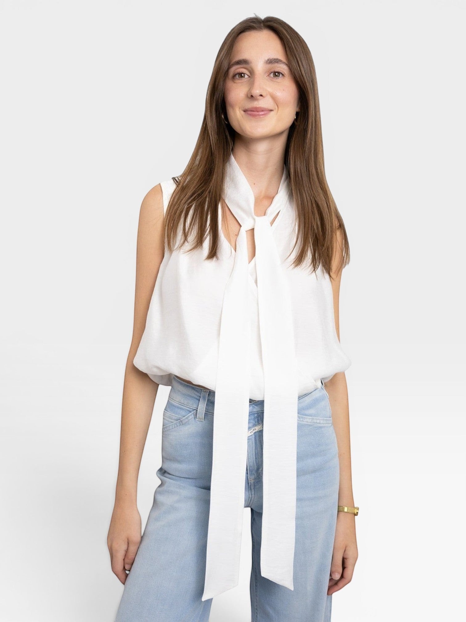 JONES + CO white sleeveless blouse with tie-neck detail, paired with light wash jeans. Model is wearing a gold bracelet.