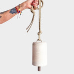 JONES + CO white stoneware bell with a star detail, beveled wood knocker, and hemp rope held by a tattooed hand.