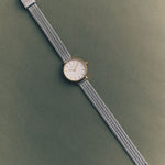 JONES + CO Beverly mesh watch in silver with a round white dial and minimalist markers on a dark green background.