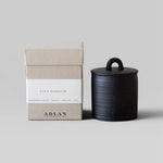 JONES + CO Eve's Nightcap petite candle in a black stoneware vessel with a matching lid next to its beige box.