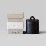 JONES + CO Eve's Nightcap petite candle in a black stoneware vessel with a matching lid next to its beige box.