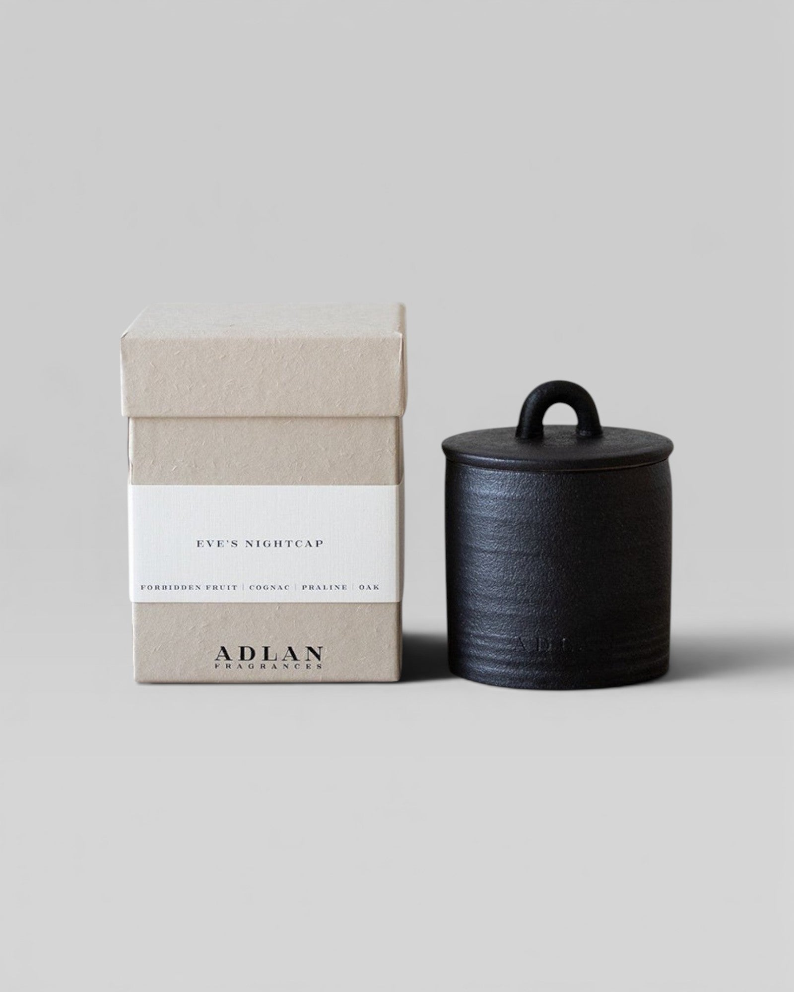 JONES + CO Eve's Nightcap petite candle in a black stoneware vessel with a matching lid next to its beige box.