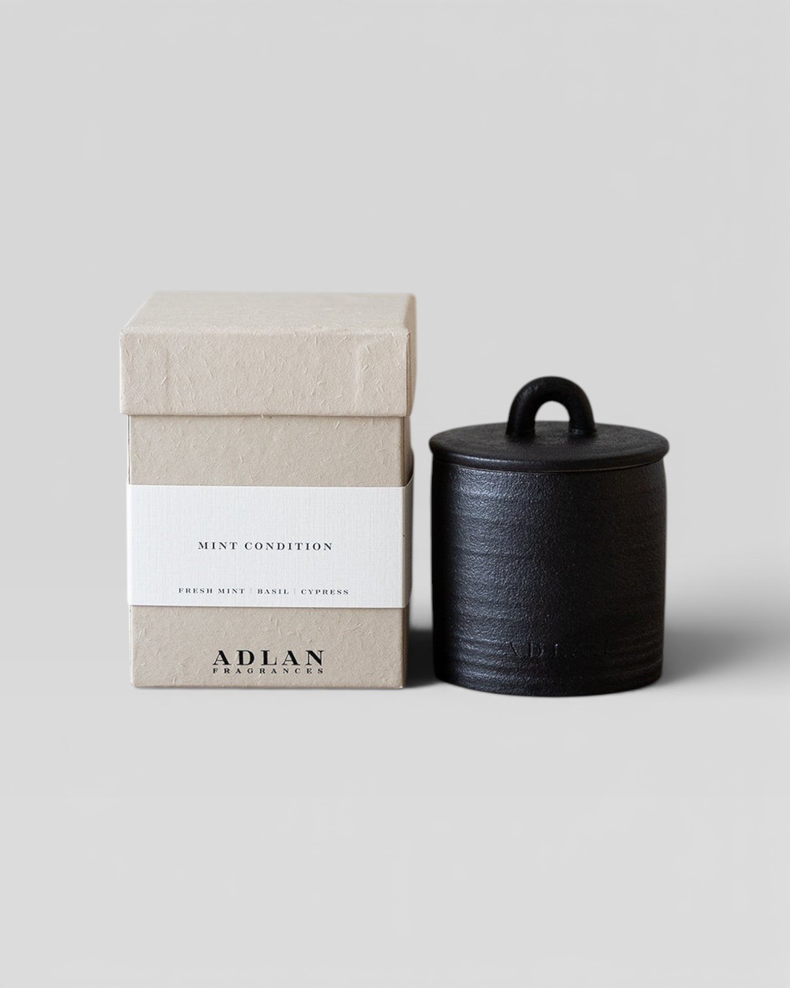 JONES + CO Mint Condition petite candle in a black stoneware vessel with a matching lid next to its beige gift box.