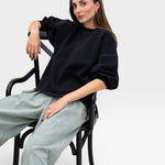 JONES + CO charcoal Fond sweatshirt with drop shoulders and raw edge hem worn by a model sitting on a black chair.