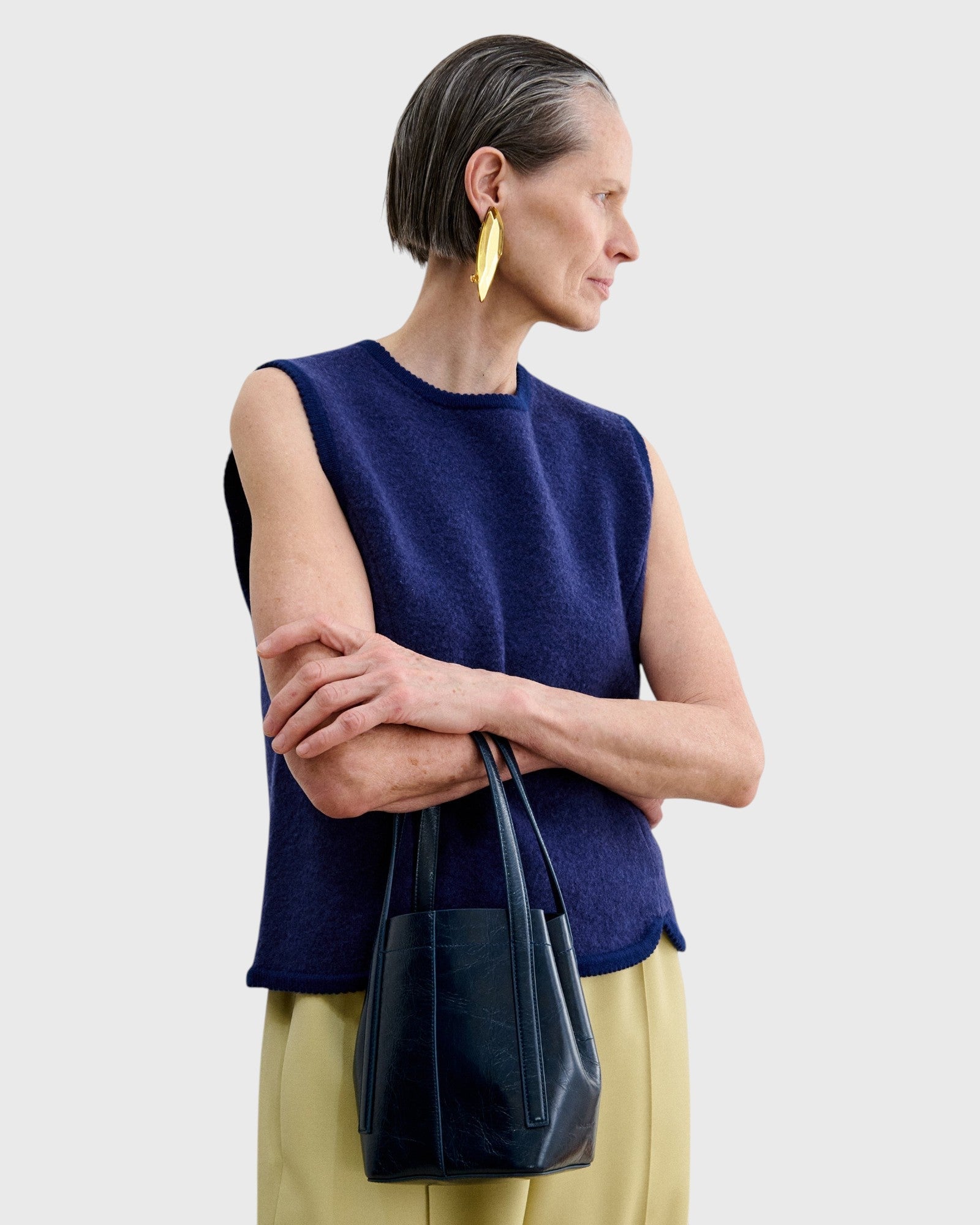 JONES + CO dark indigo lustre leather bucket bag with vertical paneling held by a woman in a navy blue knit vest.