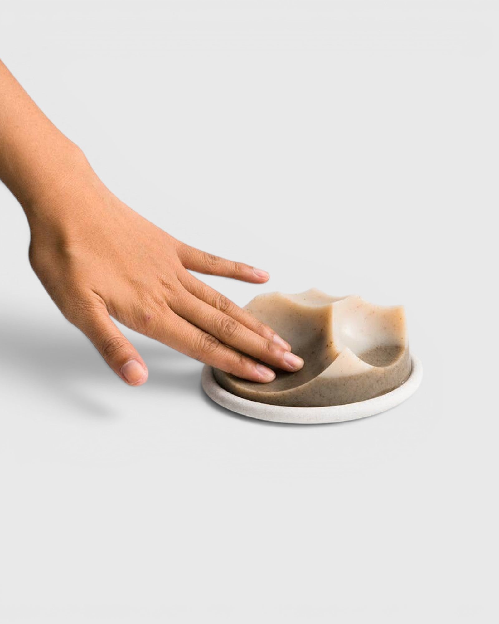 JONES + CO Erode Cardamom Vetiver soap with a unique sculpted shape resting on a white ceramic plate, touched by a hand.