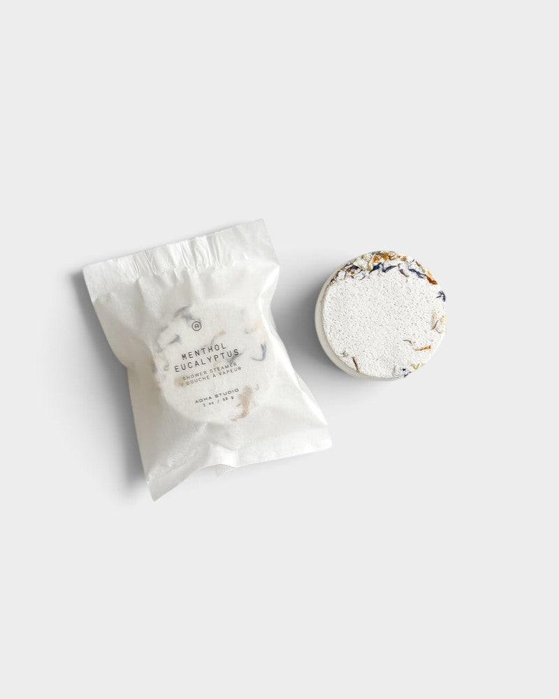 JONES + CO Menthol Eucalyptus shower steamer tablet with dried botanical petals next to its white paper packaging.