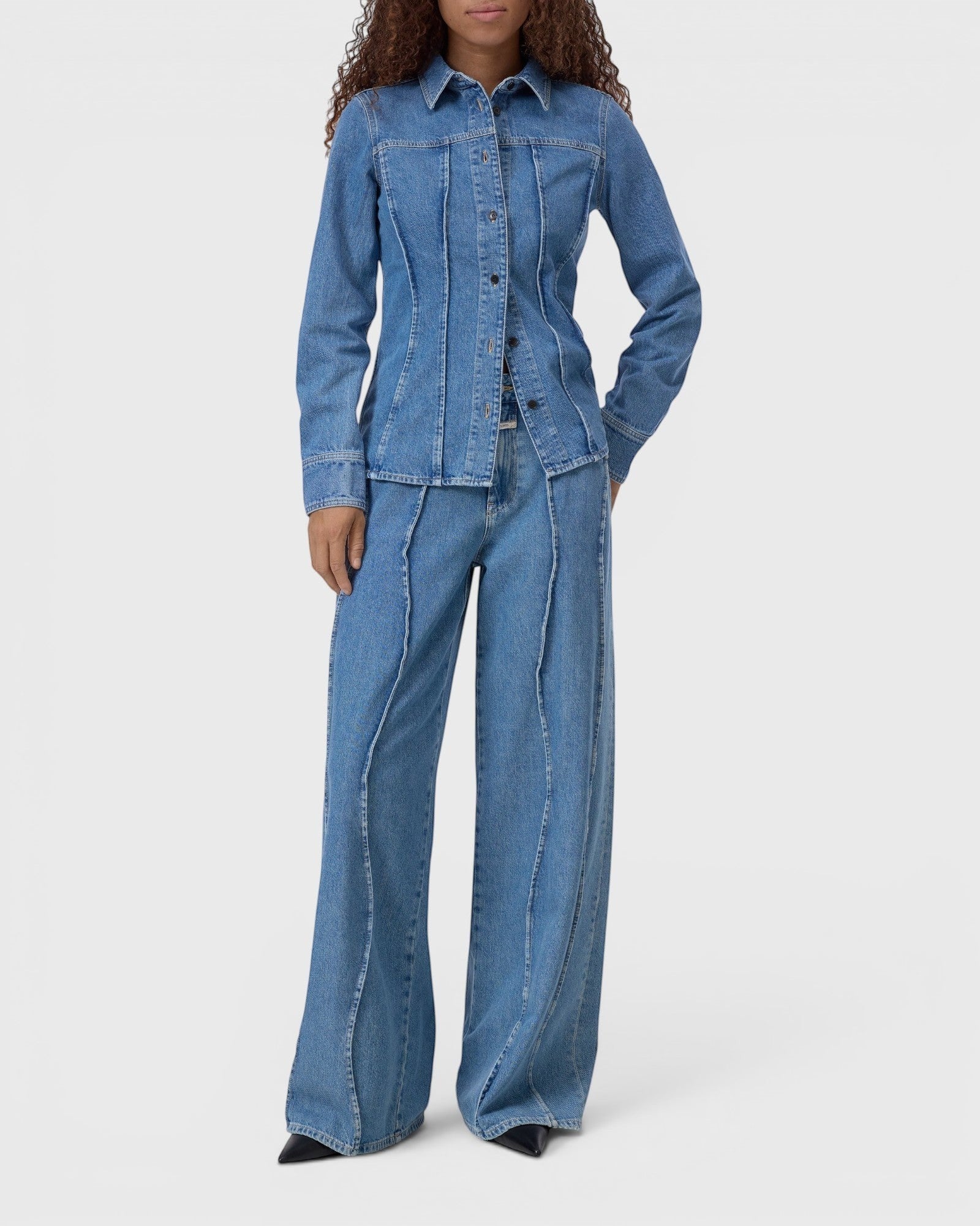 Model wearing a JONES + CO mid-blue denim shirt with curved seams, pointed collar, and button-front closure.