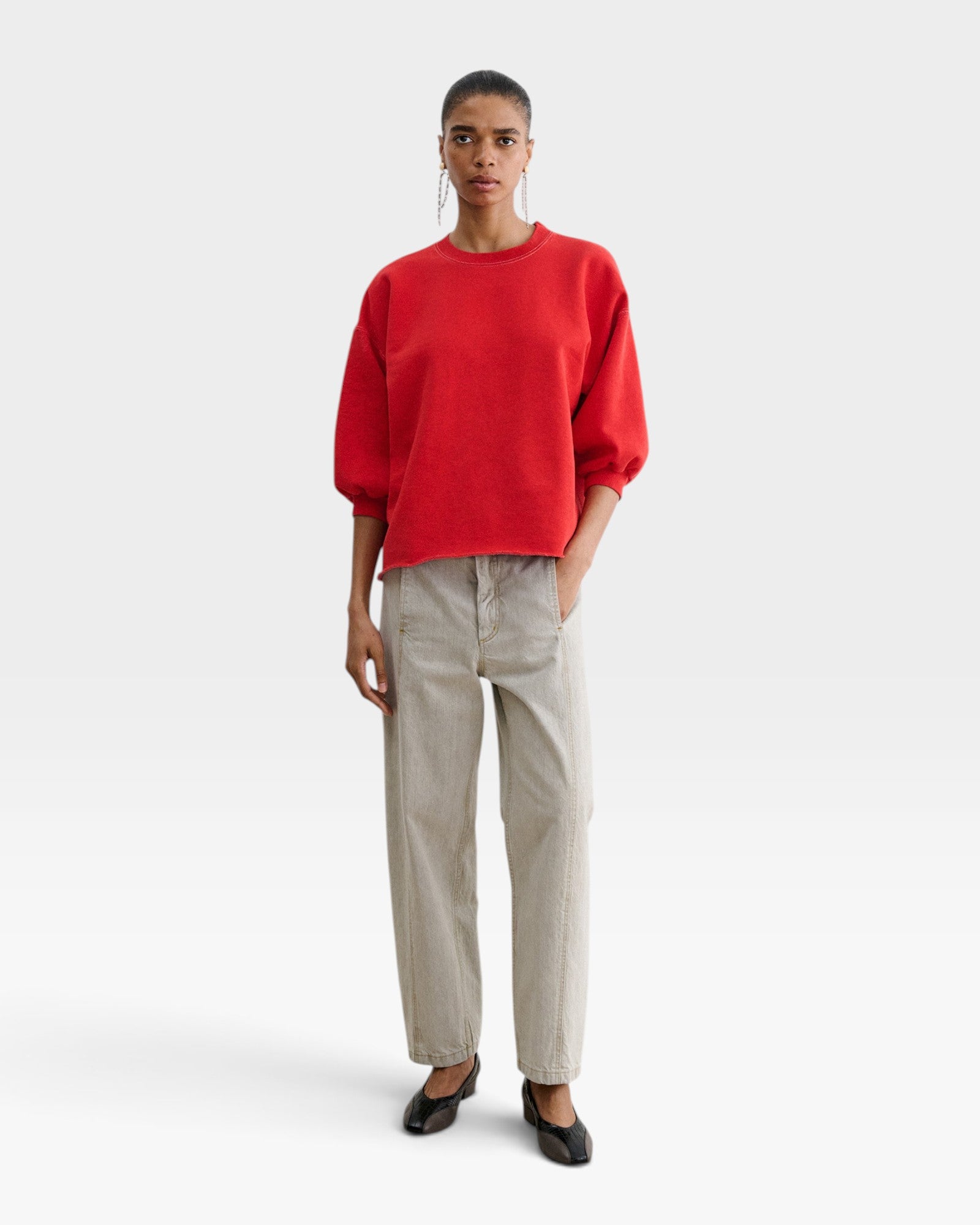 Model wearing a red JONES + CO Fond crewneck sweatshirt with drop shoulders and raw edge hem, paired with beige pants.