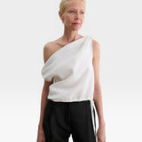 Model wearing a white JONES + CO Farfalla one-shoulder top in Italian Foam fabric with draped, gathered shoulder detail.