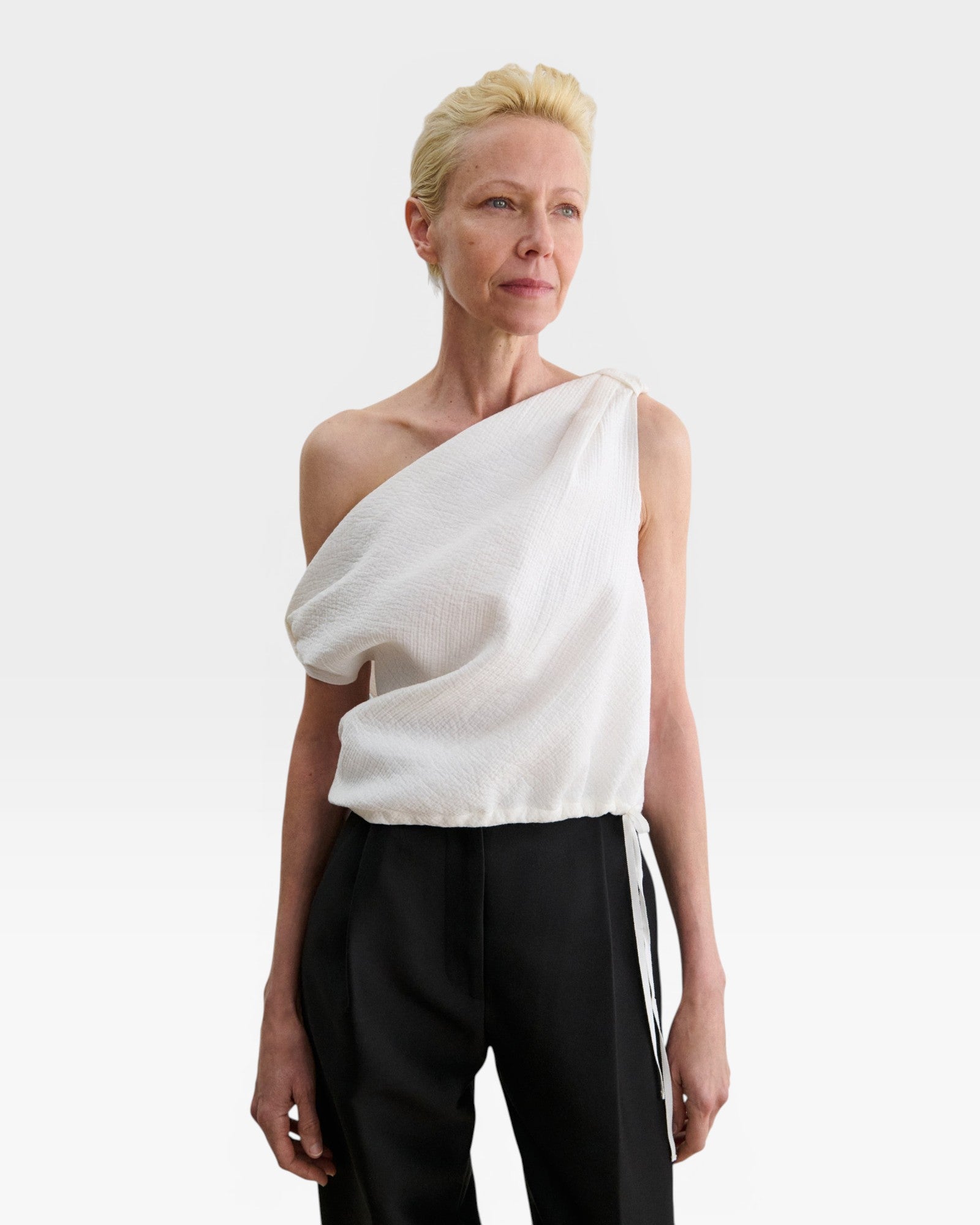 Model wearing a white JONES + CO Farfalla one-shoulder top in Italian Foam fabric with draped, gathered shoulder detail.