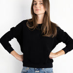 Model wearing a JONES + CO black Ziggy sweatshirt with a shrunken fit and cropped sleeves, paired with blue jeans.