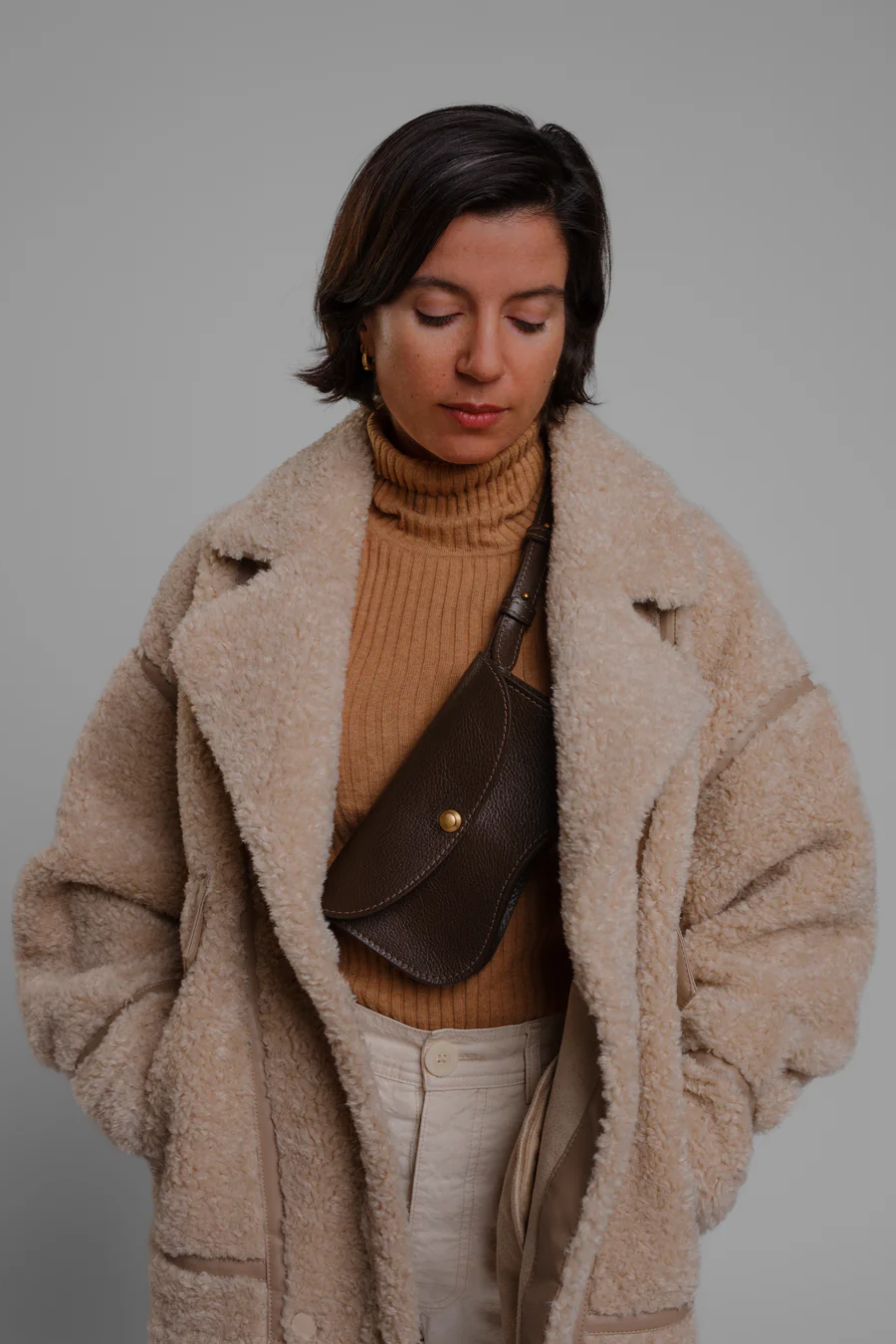 Woman wearing a beige coat and brown sweater with a brown bag against a gray background