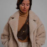 Woman wearing a beige coat and brown sweater with a brown bag against a gray background