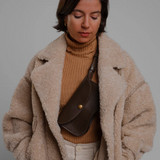 Woman wearing a beige coat and brown sweater with a brown bag against a gray background