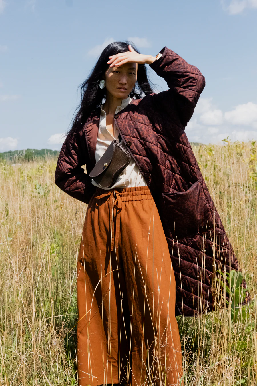 Woman in a long quilted brown coat and orange pants standing in a field with a blue sky.