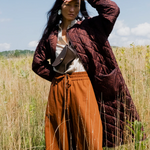 Woman in a long quilted brown coat and orange pants standing in a field with a blue sky.