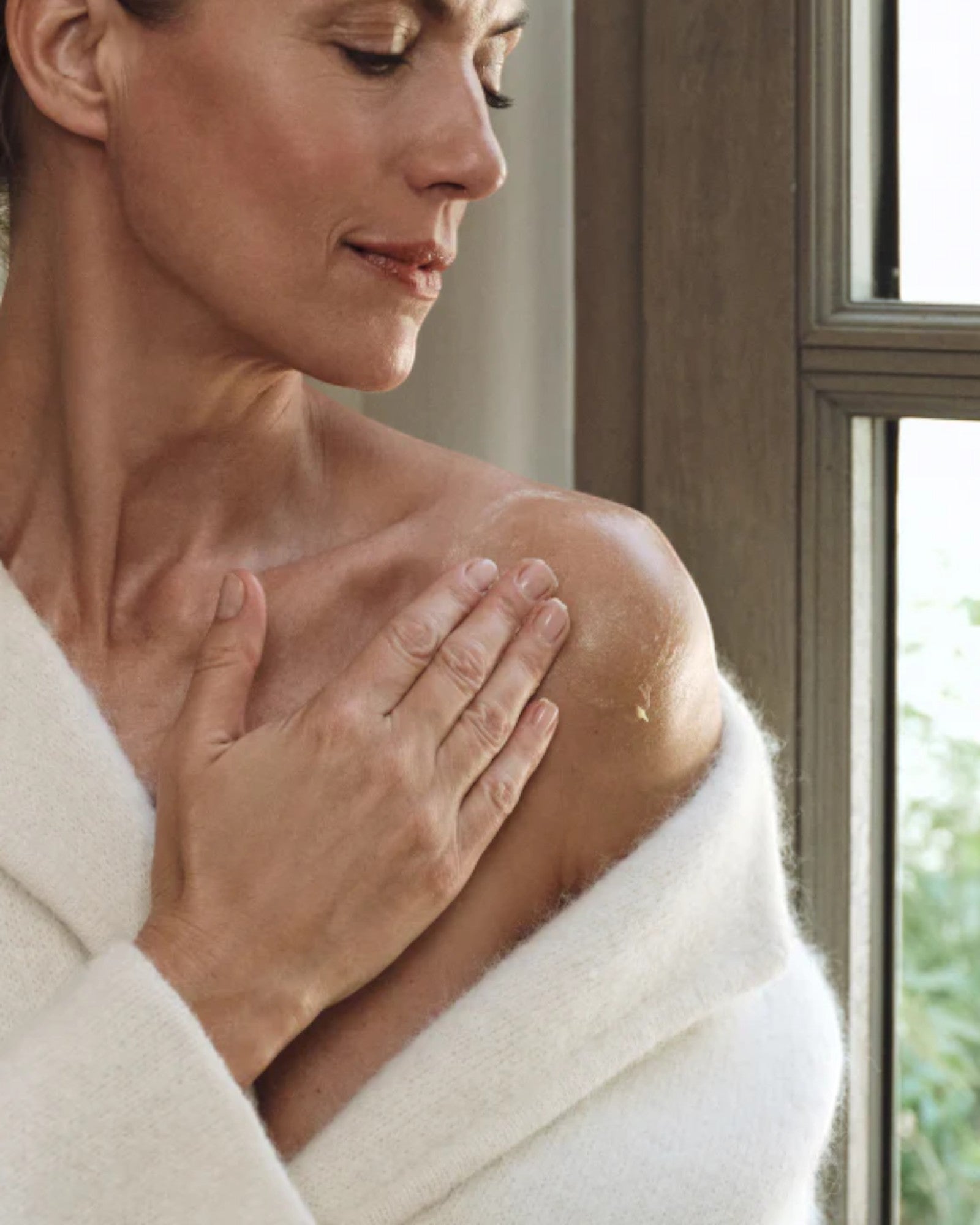 Woman in a white robe rubbing in the Oak Essentials Dew Body Butter with a window in the background