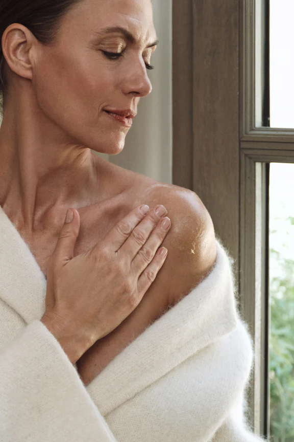 Woman in a white robe rubbing in the Oak Essentials Dew Body Butter with a window in the background