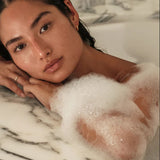 Woman in a bathtub with bubbles, leaning against a marble wall.