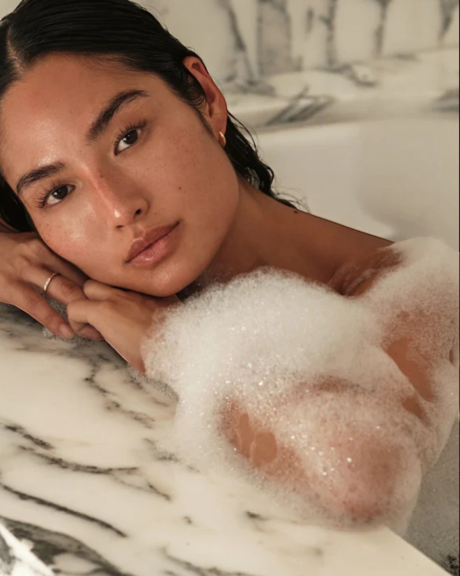 Woman in a bathtub with bubbles, leaning against a marble wall.