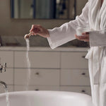 Person in a white robe standing by a sink, pouring a white powder into running water.