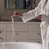 Person in a white robe standing by a sink, pouring a white powder into running water.