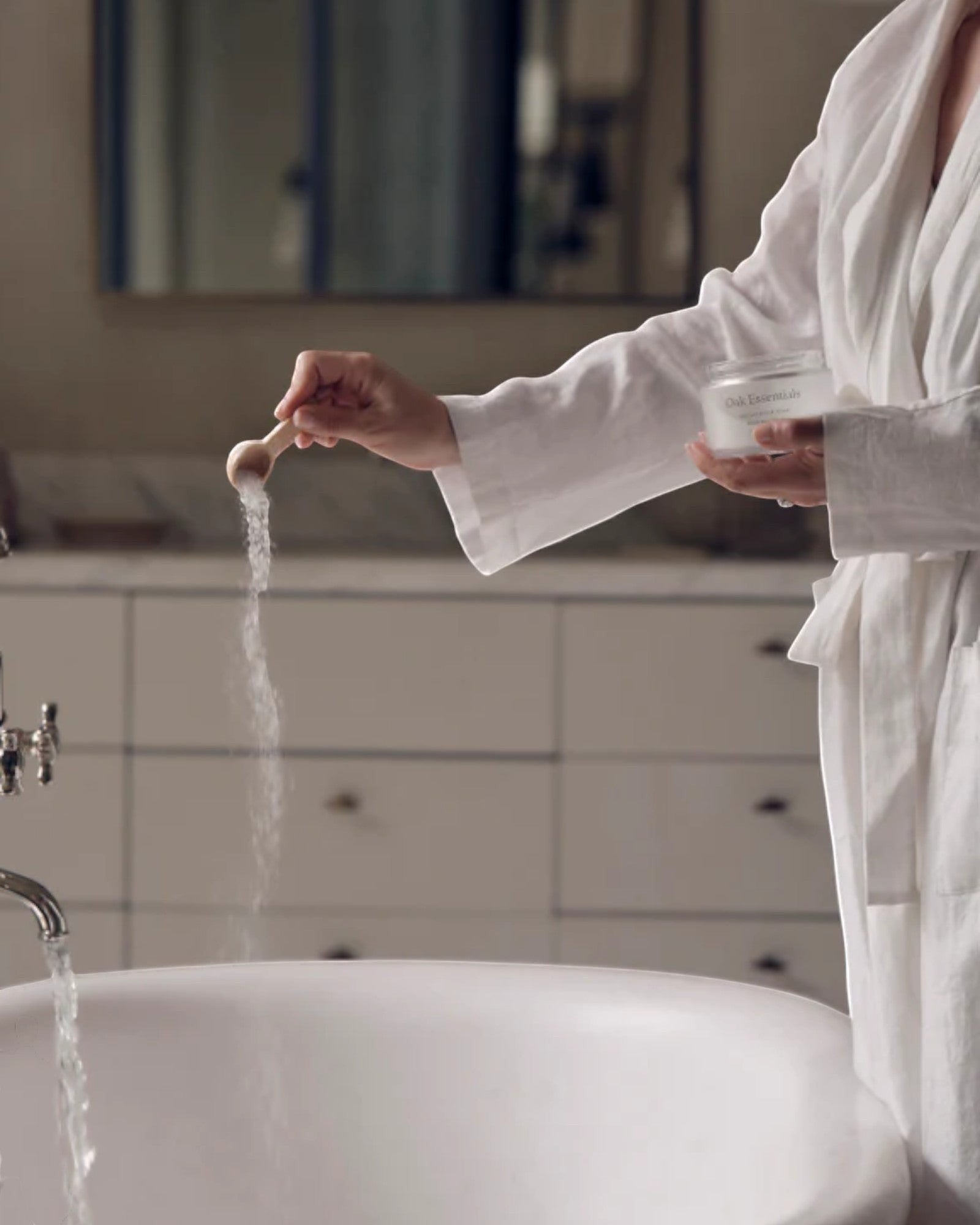 Person in a white robe standing by a sink, pouring a white powder into running water.