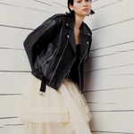 Person wearing a black leather jacket and layered outfit against a white wooden wall.