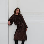 Woman wearing a dark brown sequin top and matching skirt against a white wall.