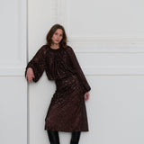 Woman wearing a dark brown sequin top and matching skirt against a white wall.