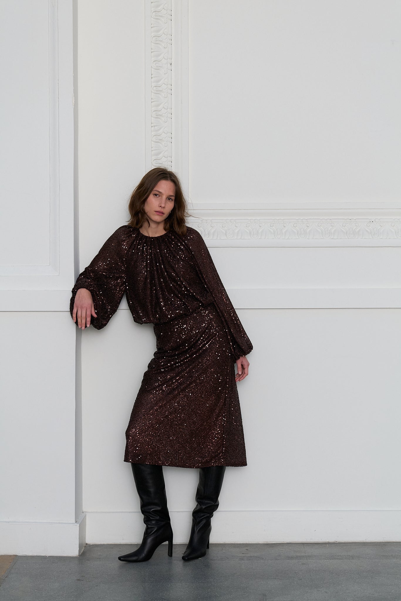 Woman wearing a dark brown sequin top and matching skirt against a white wall.
