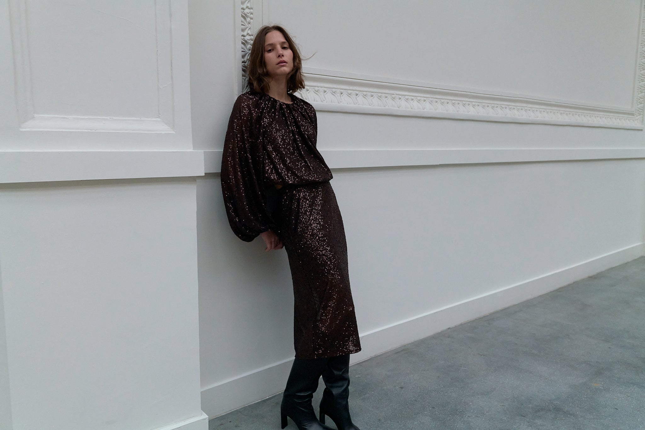 Woman in a brown sequin dress standing against a white wall.