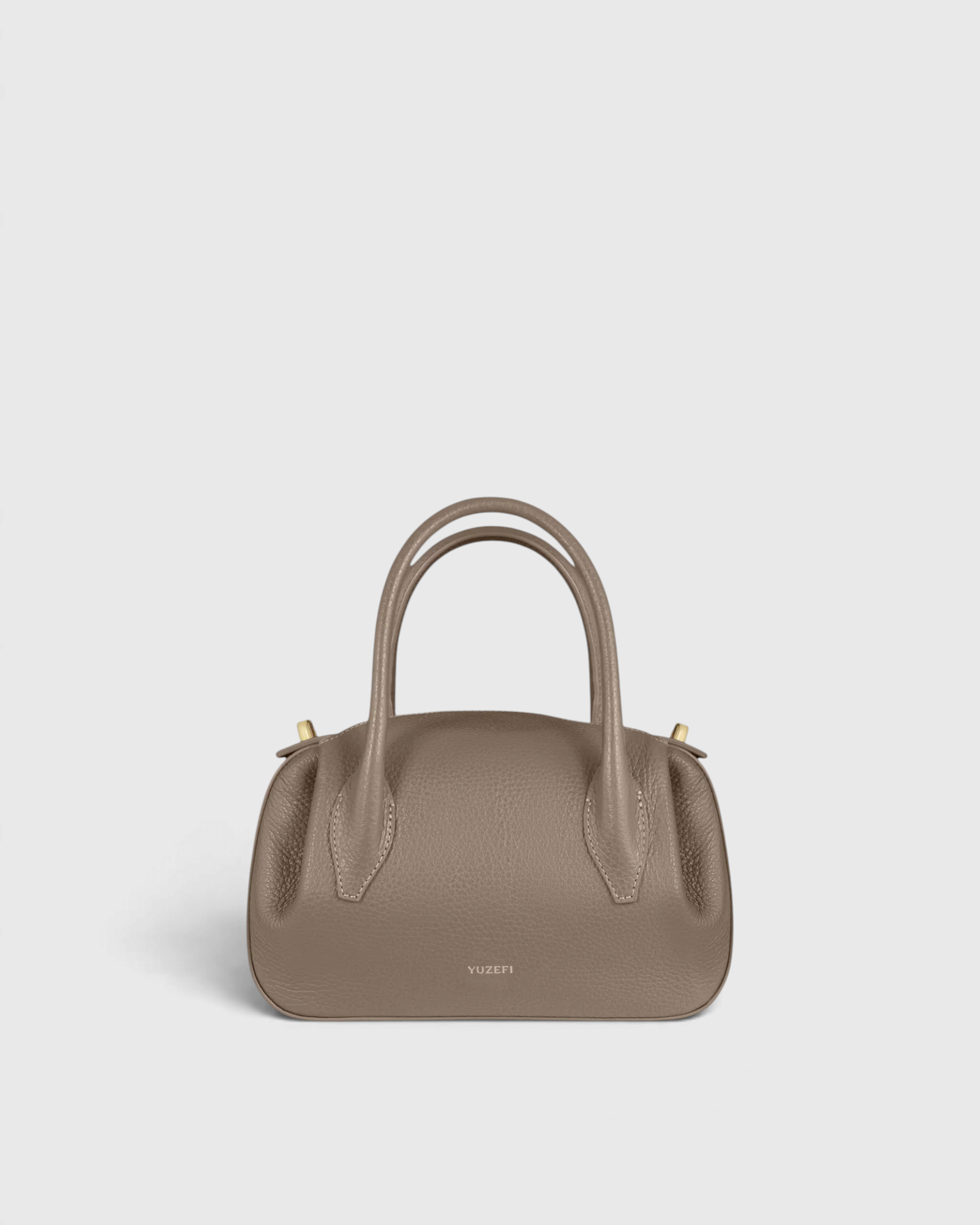 JONES + CO Oyster Mini Duffle in taupe pebble grained Italian leather with gold-tone hardware and top handles.