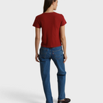 A woman is shown from behind wearing a red short-sleeve t-shirt and blue jeans, standing against a draped beige fabric backdrop.