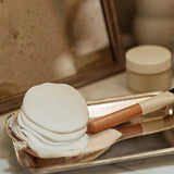White bath bombs, wooden scrubber, and brush on a gold tray with a mirror and skincare products in the background.