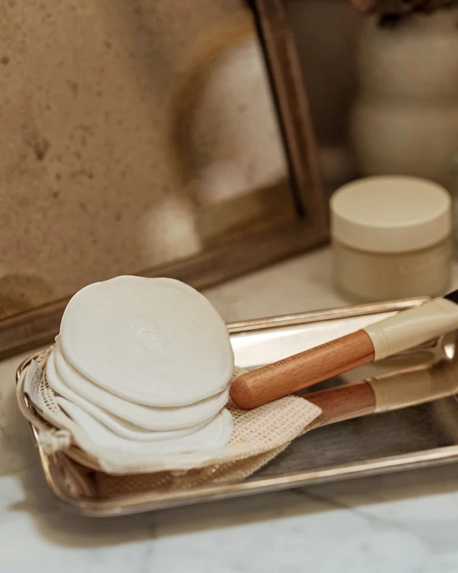 White bath bombs, wooden scrubber, and brush on a gold tray with a mirror and skincare products in the background.