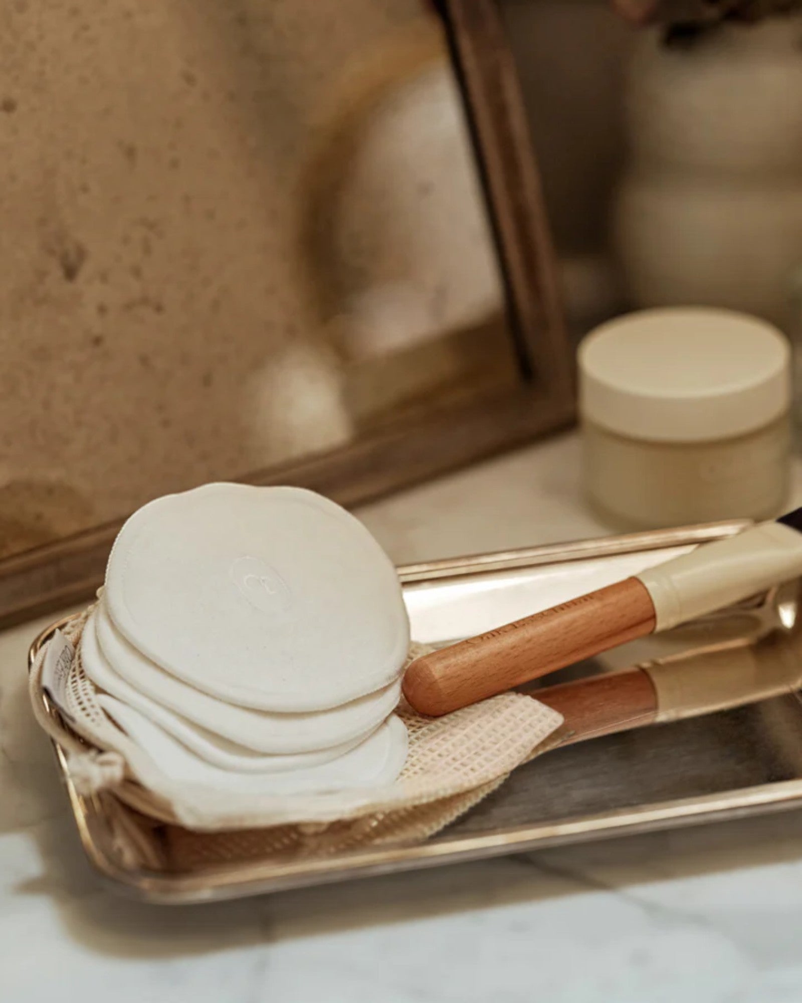 White bath bombs, wooden scrubber, and brush on a gold tray with a mirror and skincare products in the background.