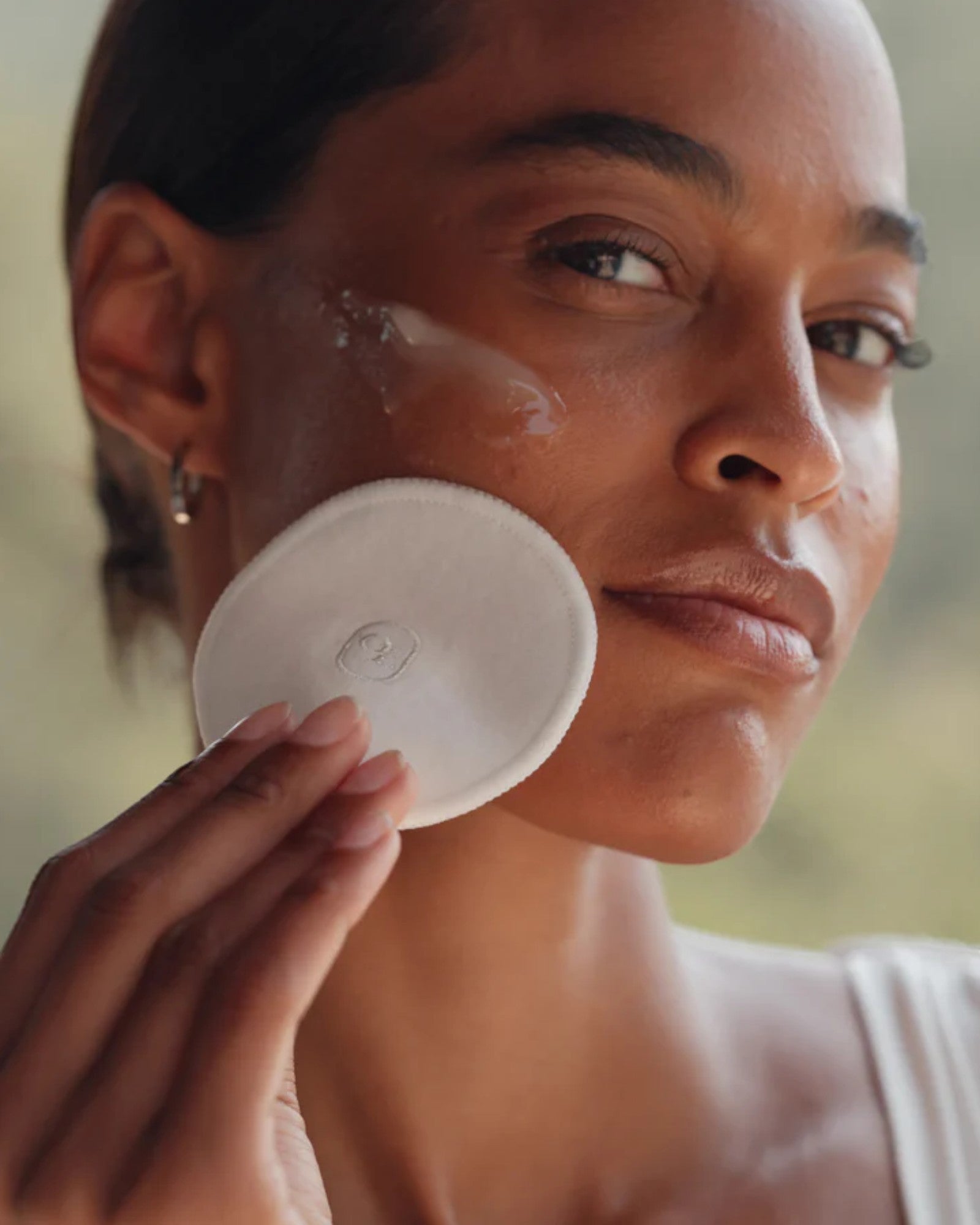 Woman applying a white round compact to her face with a blurred natural background