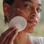 Woman applying a white round compact to her face with a blurred natural background